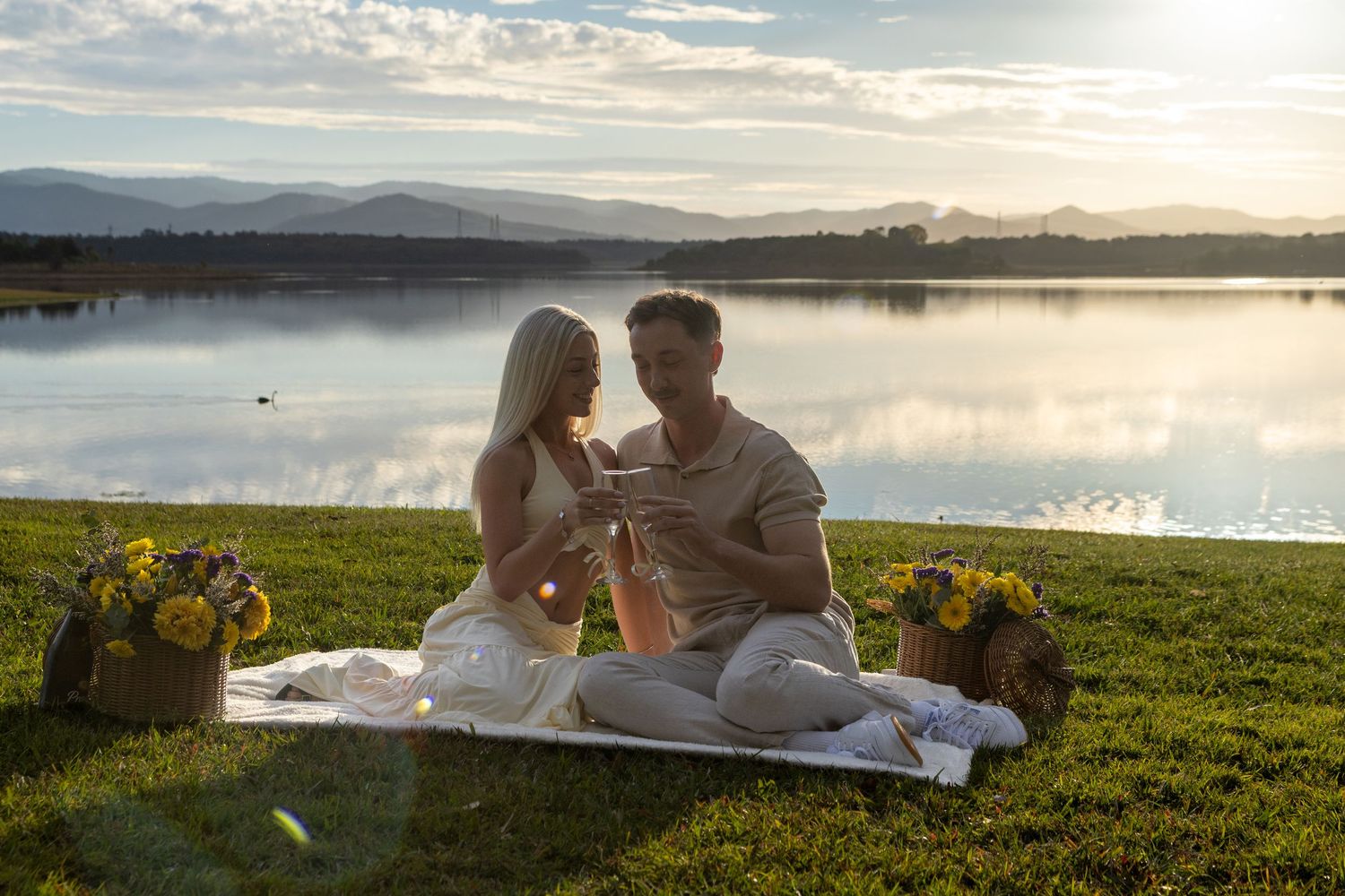 Couple sharing a romantic lakeside picnic at sunset with yellow flowers and mountain views.