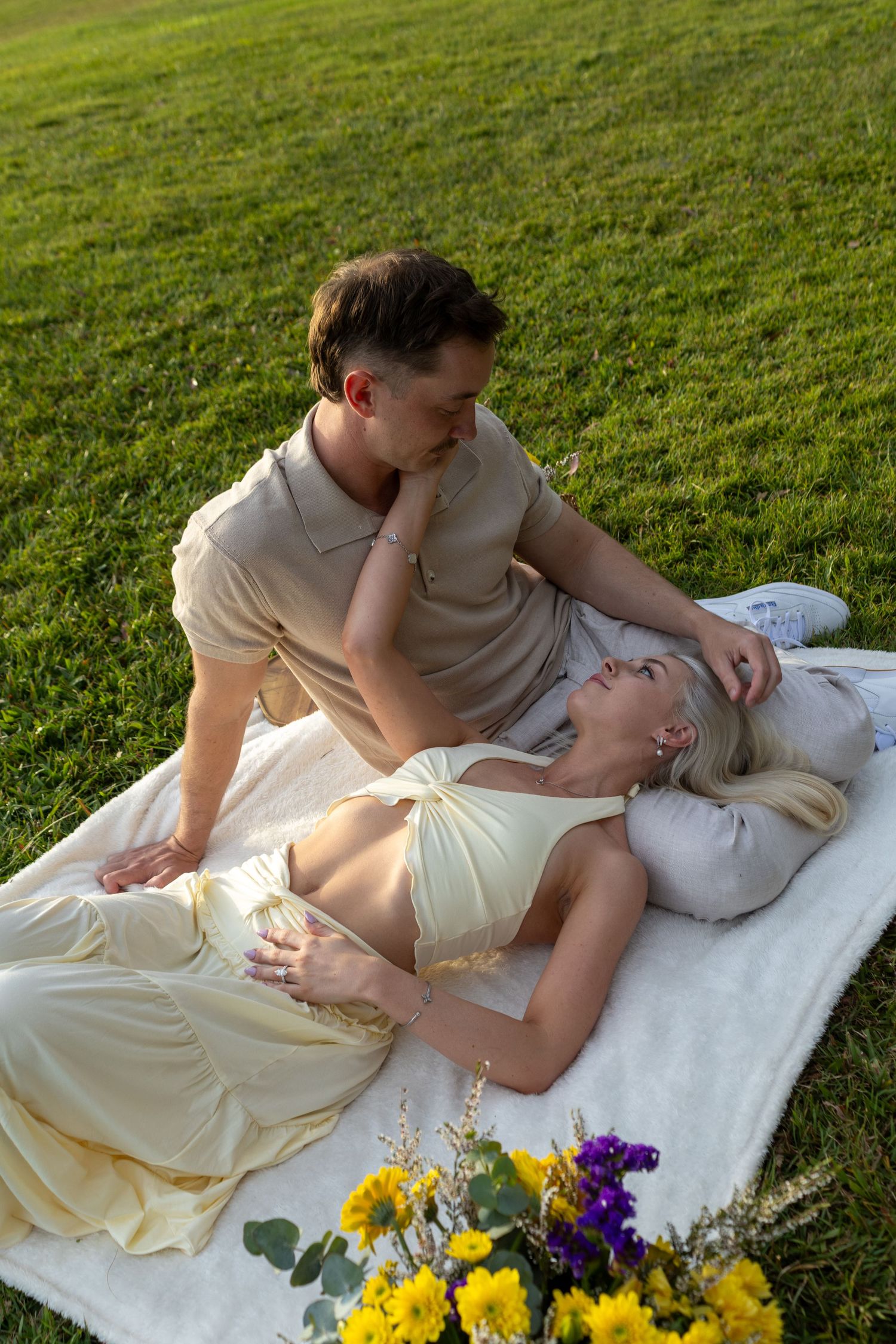 Couple sharing a romantic moment on a white blanket in lush green grass with yellow and purple flowers nearby.