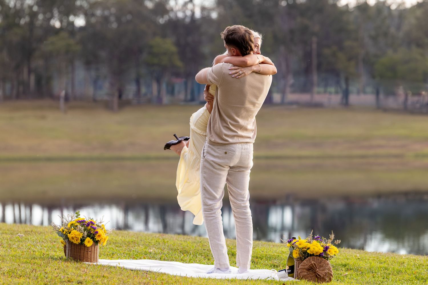 Couple embracing outdoors near a lake with yellow flower basket arrangements on green grass.