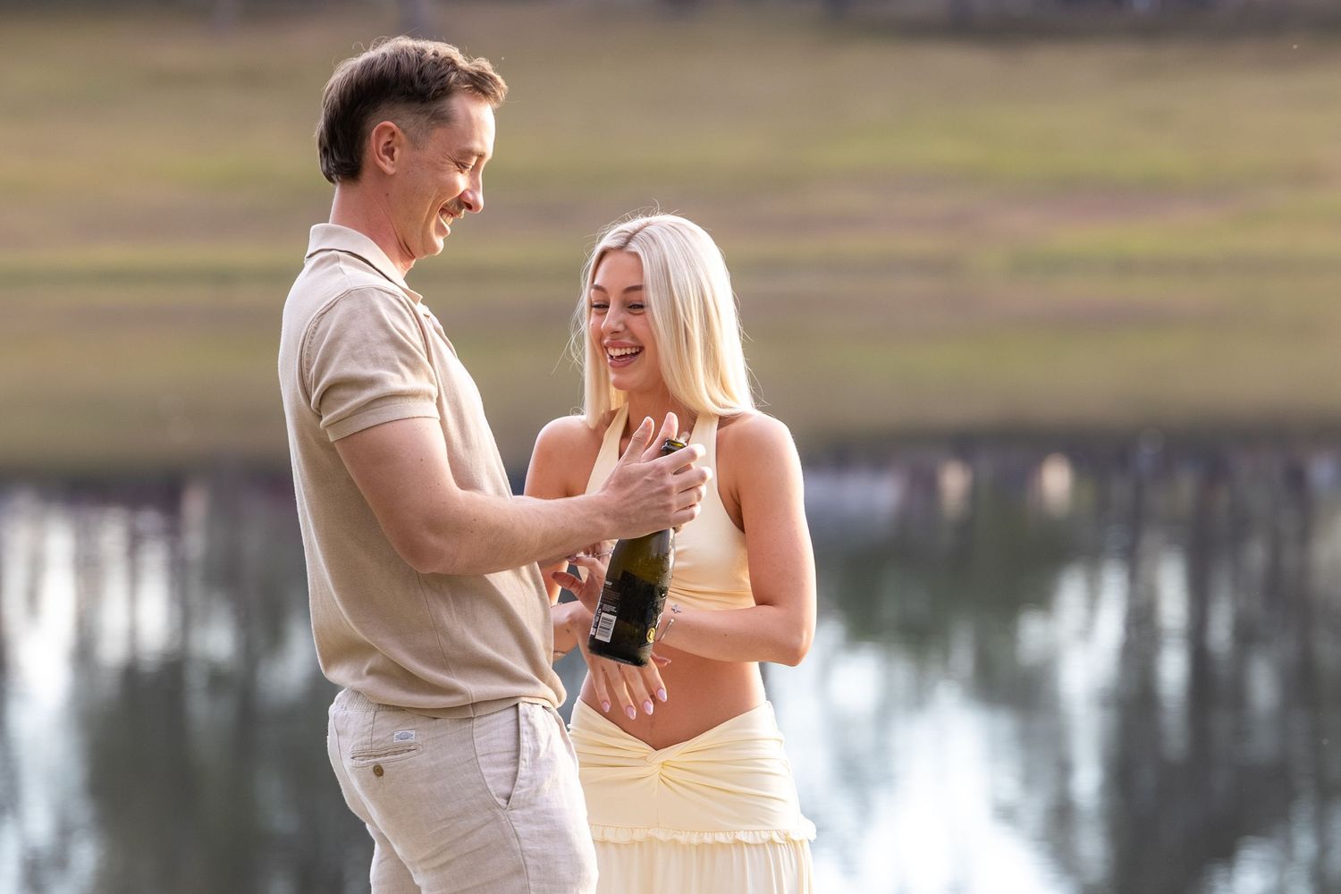 Couple sharing champagne by a lake, man in beige outfit and woman in yellow dress, celebrating outdoors.
