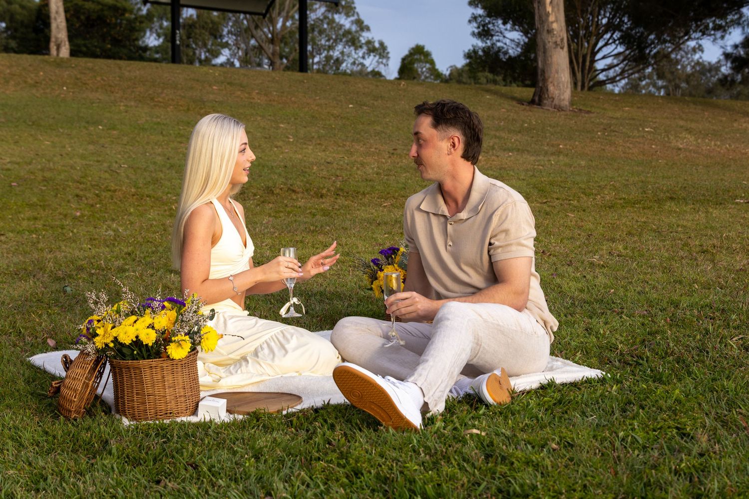 Couple enjoying a romantic picnic on green grass with a wicker basket of yellow flowers on a sunny day.