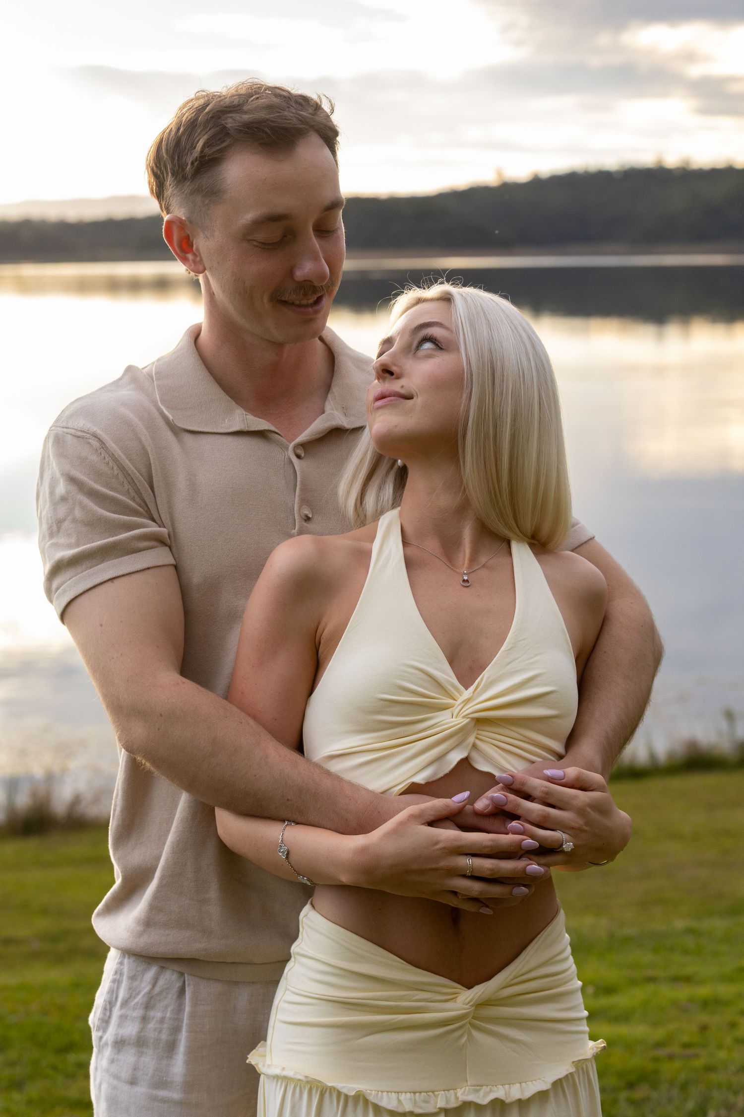 Couple embracing by a serene lake at sunset, woman in cream halter dress, man in beige polo shirt.