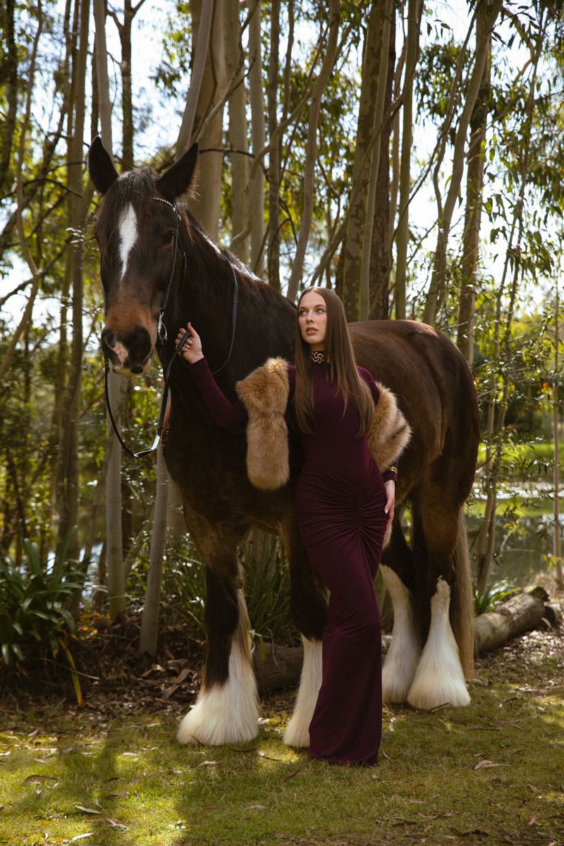 Woman in burgundy gown poses with large shire horse in a serene forest setting near a pond during a photoshoot in sydney with a horse