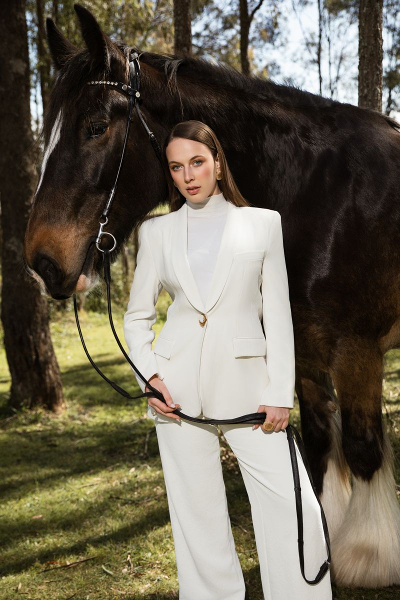 Woman in white suit holding reins beside a dark brown horse in a forest setting during a photoshoot in sydney with a horse