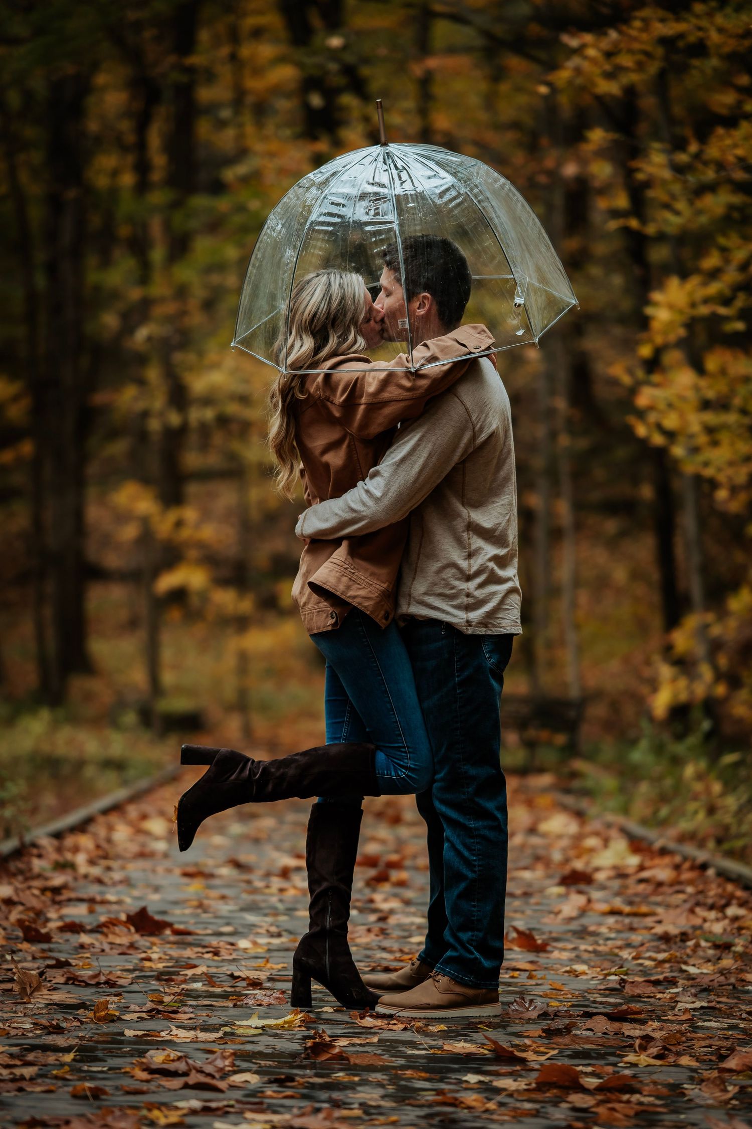 Couple kissing under clear umbrella on leaf-covered path in autumn forest, wearing jackets and boots.