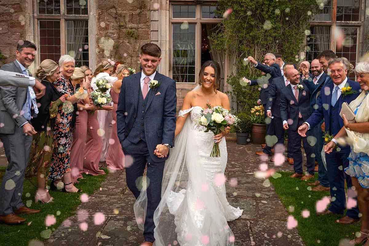 Newlywed couple walks through confetti shower surrounded by cheering wedding guests at Askham Hall wedding venue near Penrith