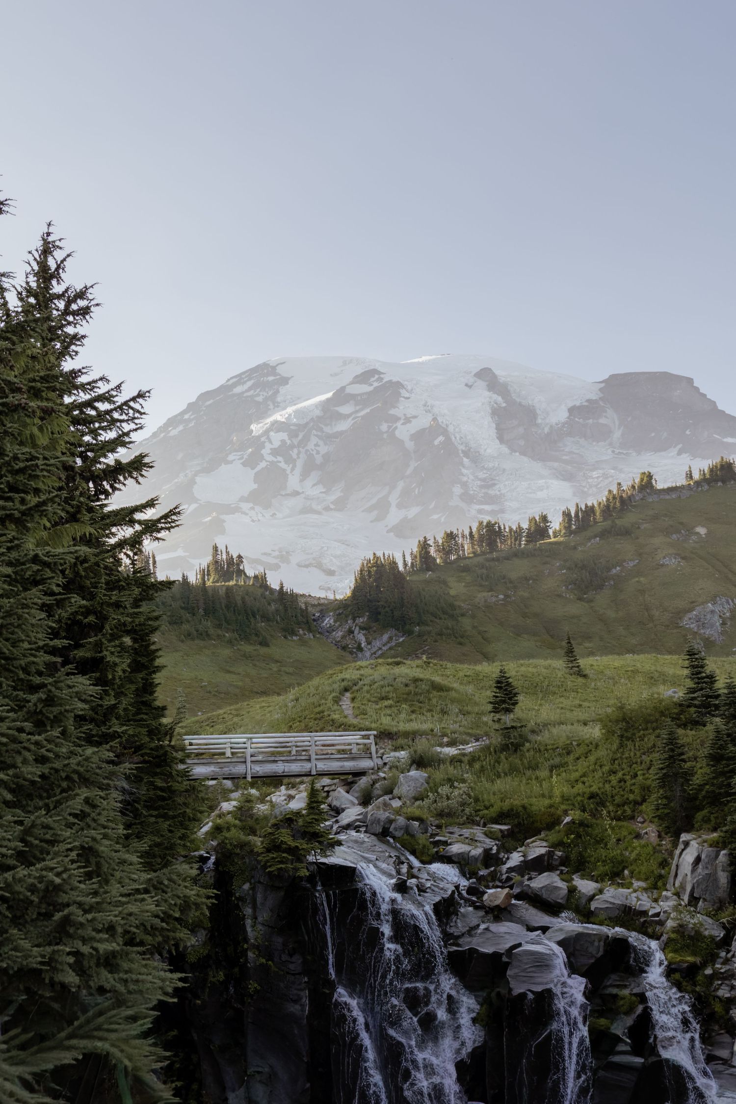 Snow-capped Mount Rainier rises above green meadows, evergreen trees, and a wooden bridge over a cascading waterfall.