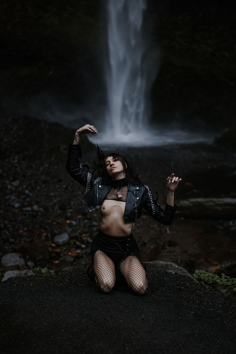 Moody fashion photoshoot featuring a model in dark clothing posing dramatically near a misty waterfall.