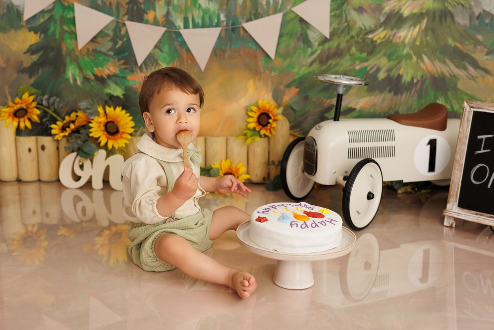 First birthday celebration scene with cake, vintage car prop and sunflower decorations against forest backdrop.