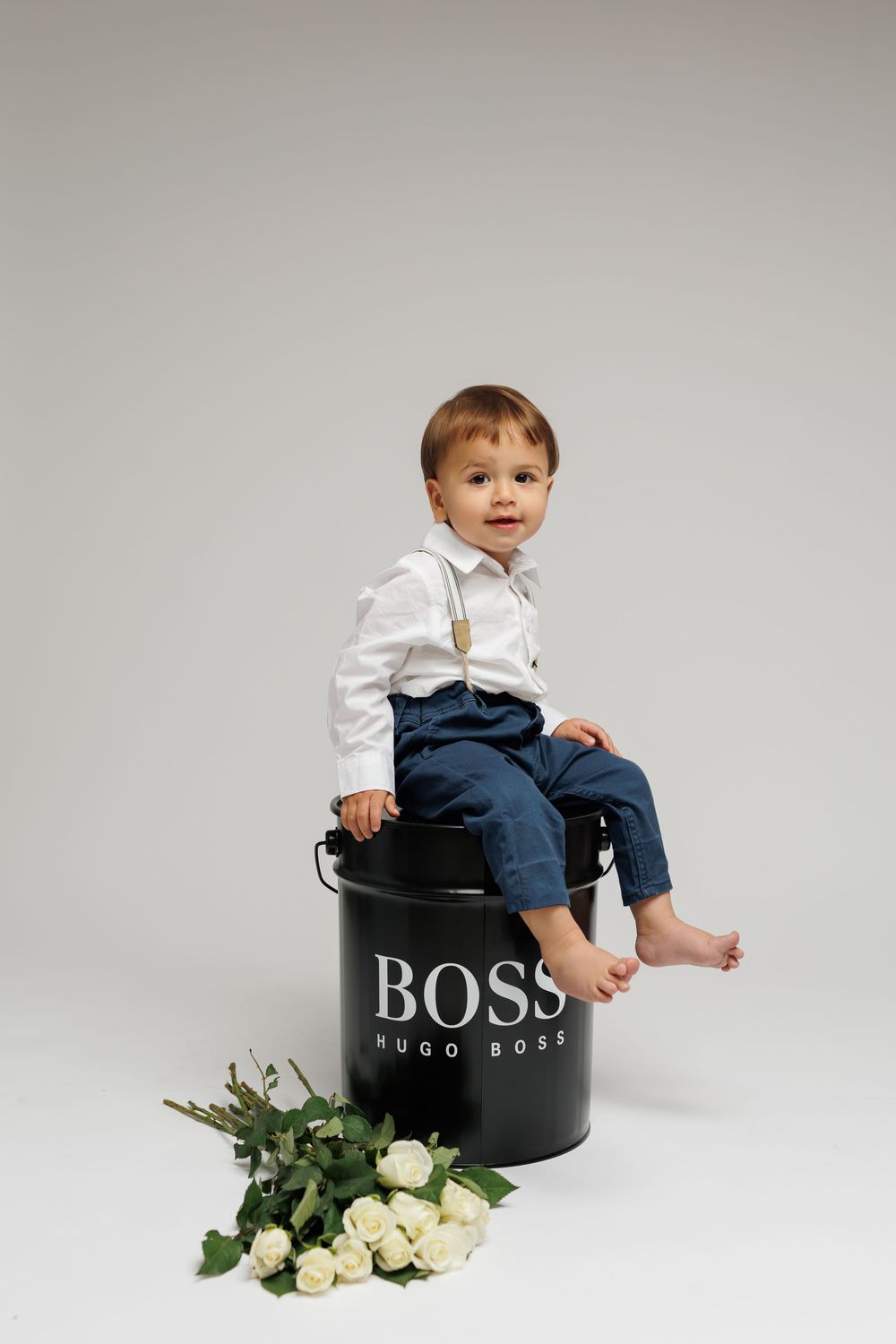 Toddler in white shirt and jeans sits on black BOSS container with white roses beside on light background.