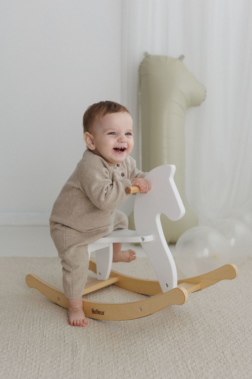 Smiling baby in beige knit romper standing at a white wooden rocking horse toy in a bright nursery room.