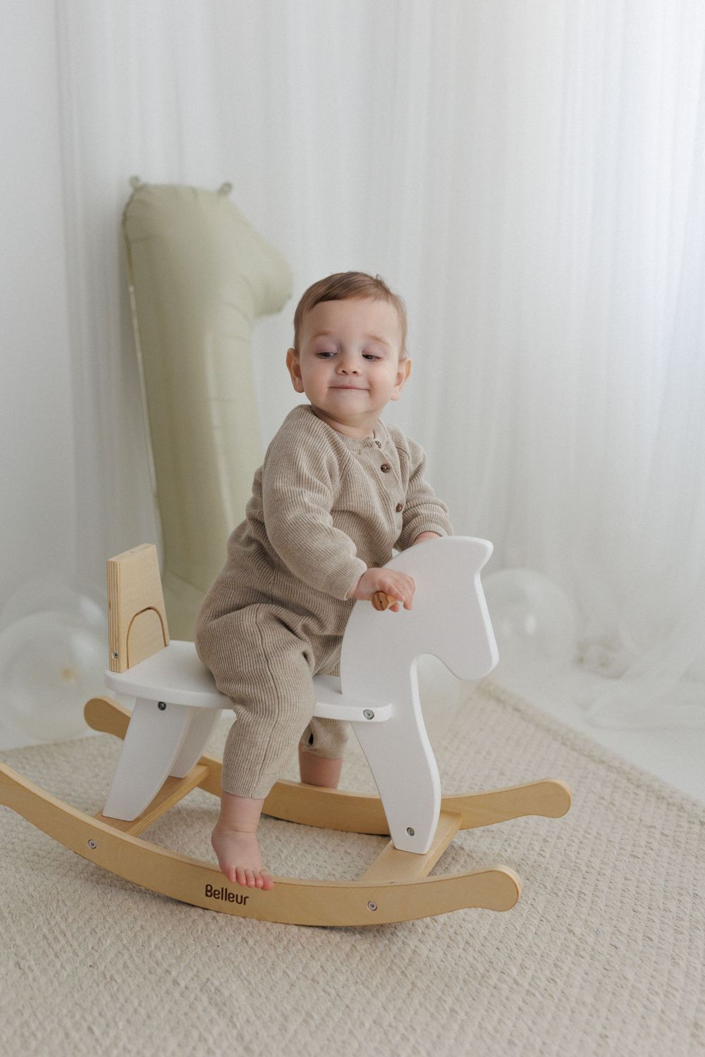 Smiling baby in beige romper riding a white and wood Bofleur rocking horse toy in a bright nursery room.