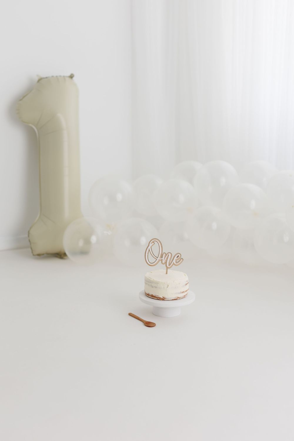White first birthday cake smash setup with number one balloon, white balloons, and cake topper reading One.