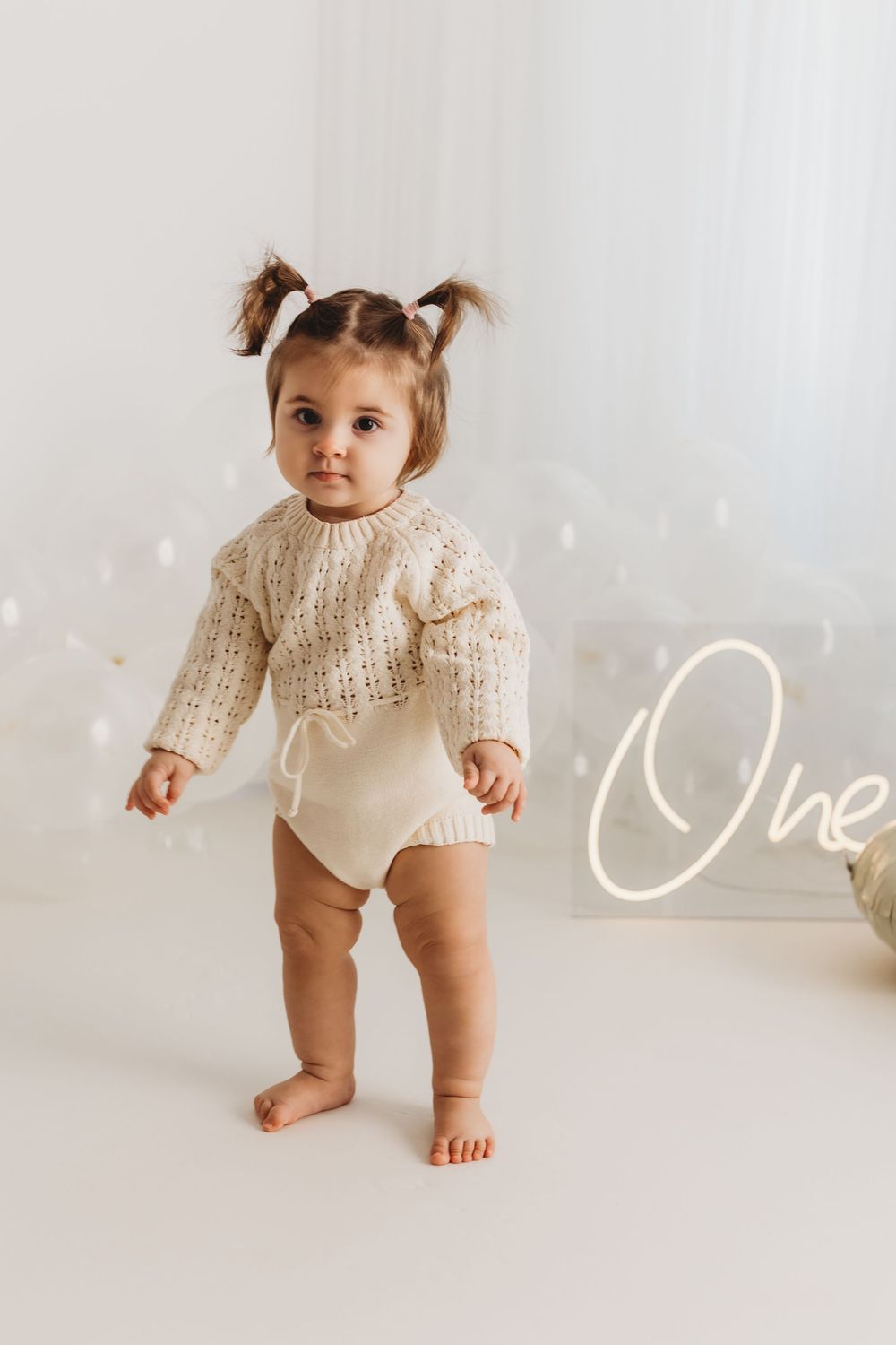 Adorable baby girl in cream knit sweater romper standing barefoot near a glowing One neon sign for first birthday.