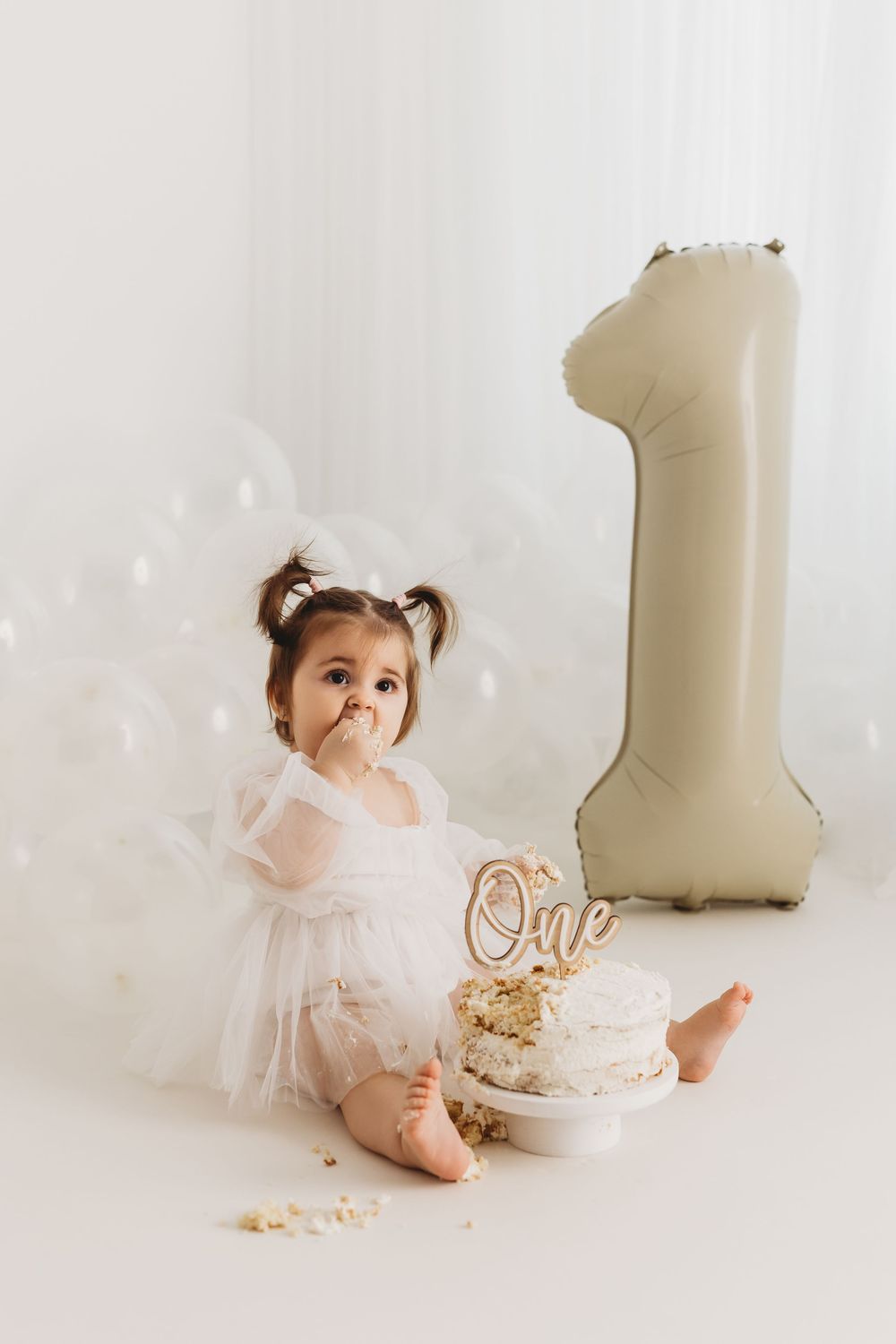 Baby girl in white tutu dress smashing first birthday cake beside a large number 1 balloon at cake smash photoshoot.