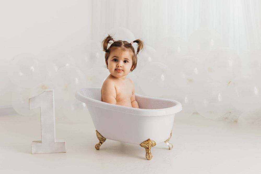 Cute baby girl sitting in a small white clawfoot bathtub with gold feet, next to a number one sign.