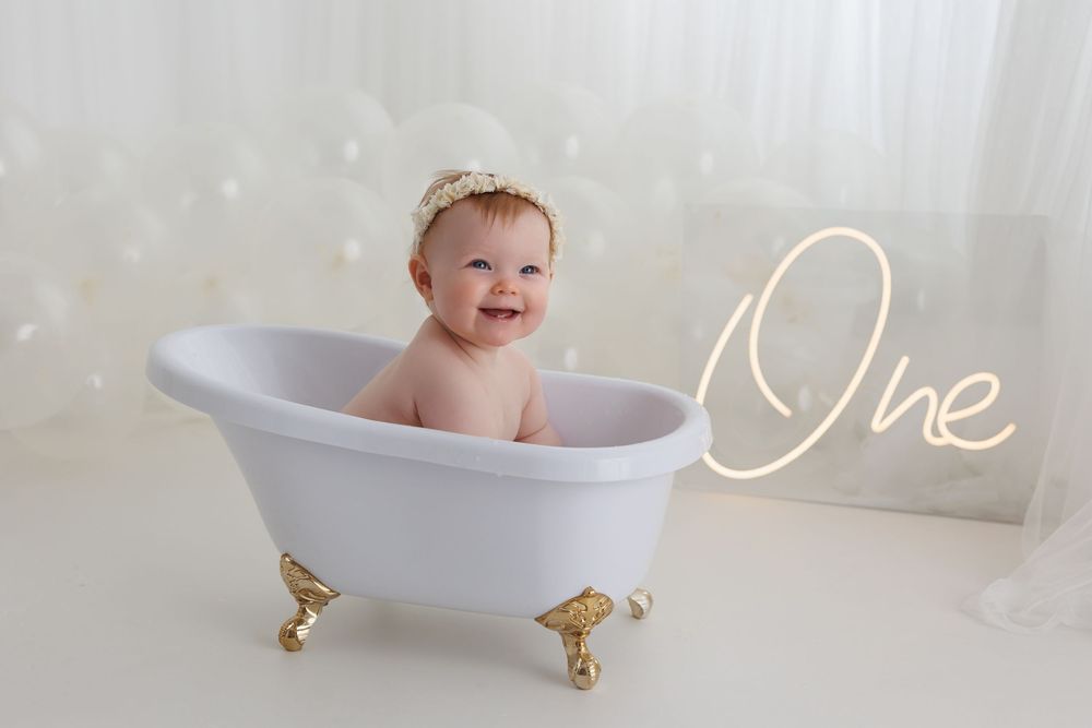 Smiling baby sitting in a white mini clawfoot bathtub beside a glowing One neon sign for a first birthday photo.