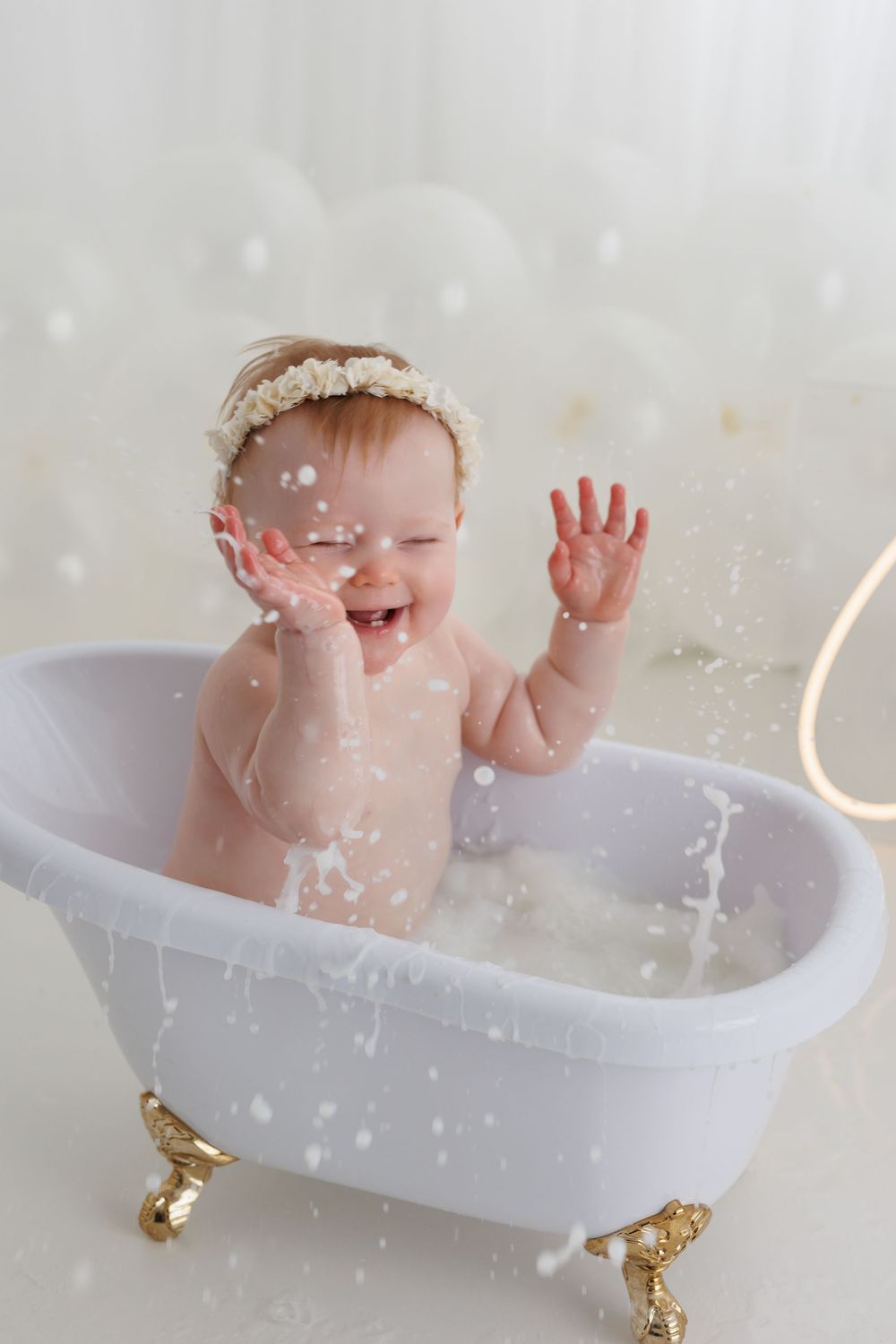 Laughing baby girl with floral headband splashing in a small white clawfoot bathtub with gold feet.