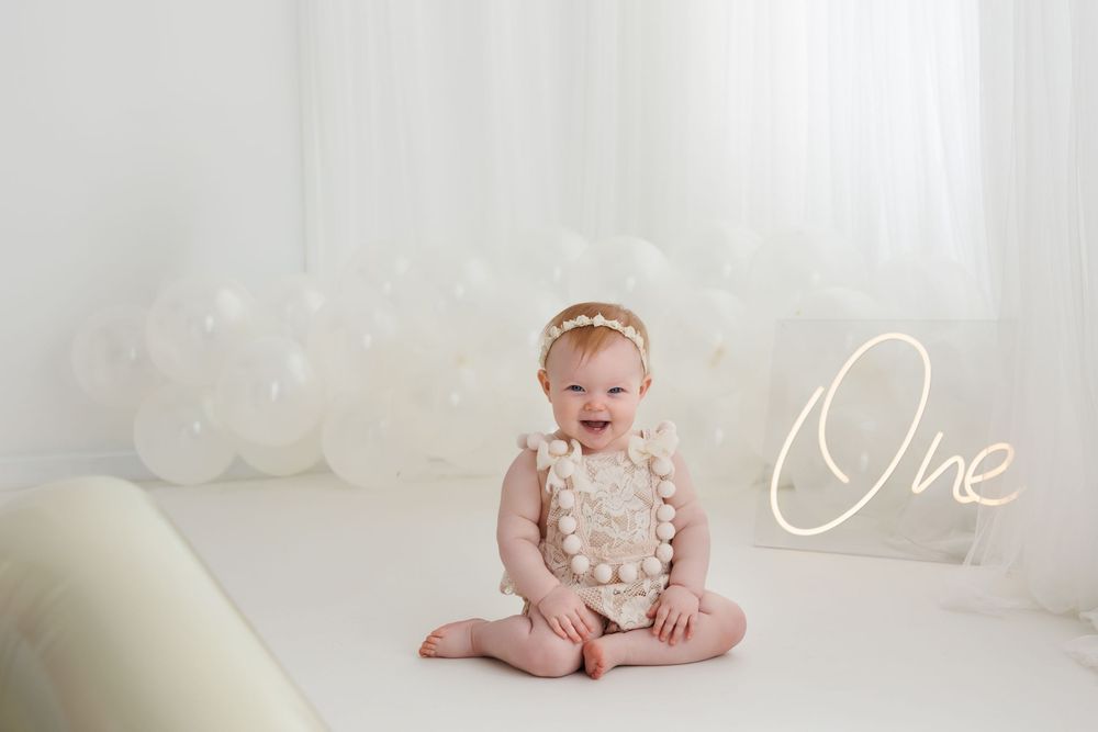 A smiling baby girl in a white romper sits before white balloons with a glowing “One” sign for her first birthday.