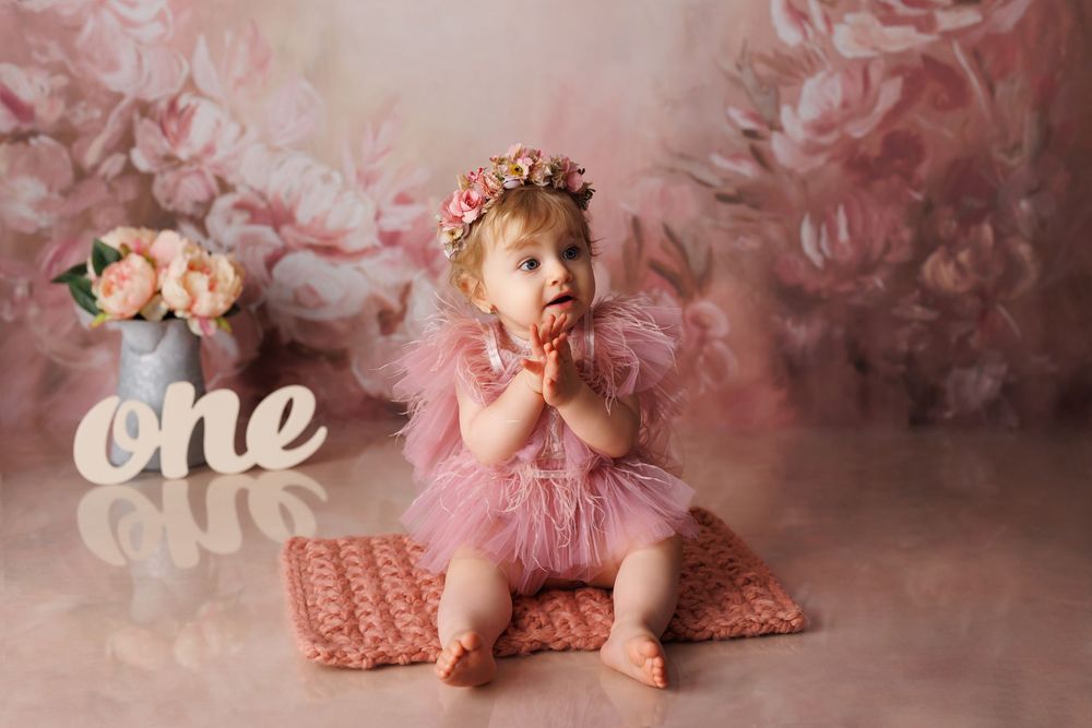 A baby in a pink tutu and floral crown sits on a cushion next to decorative 'one' letters in a dreamy pink setting.