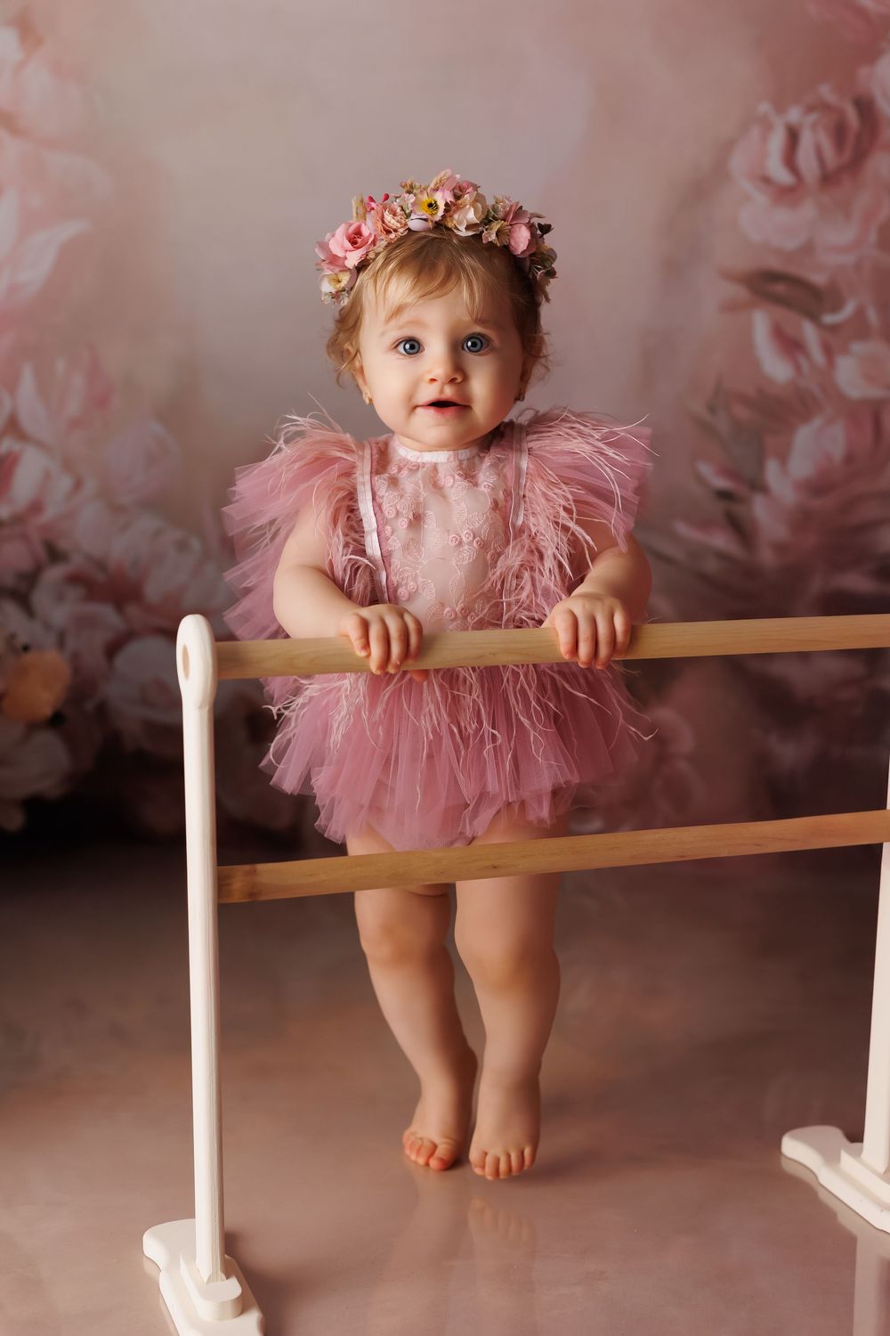 Baby wearing pink tutu dress sits on ballet barre in dreamy studio setting with soft pink smoky background effect.