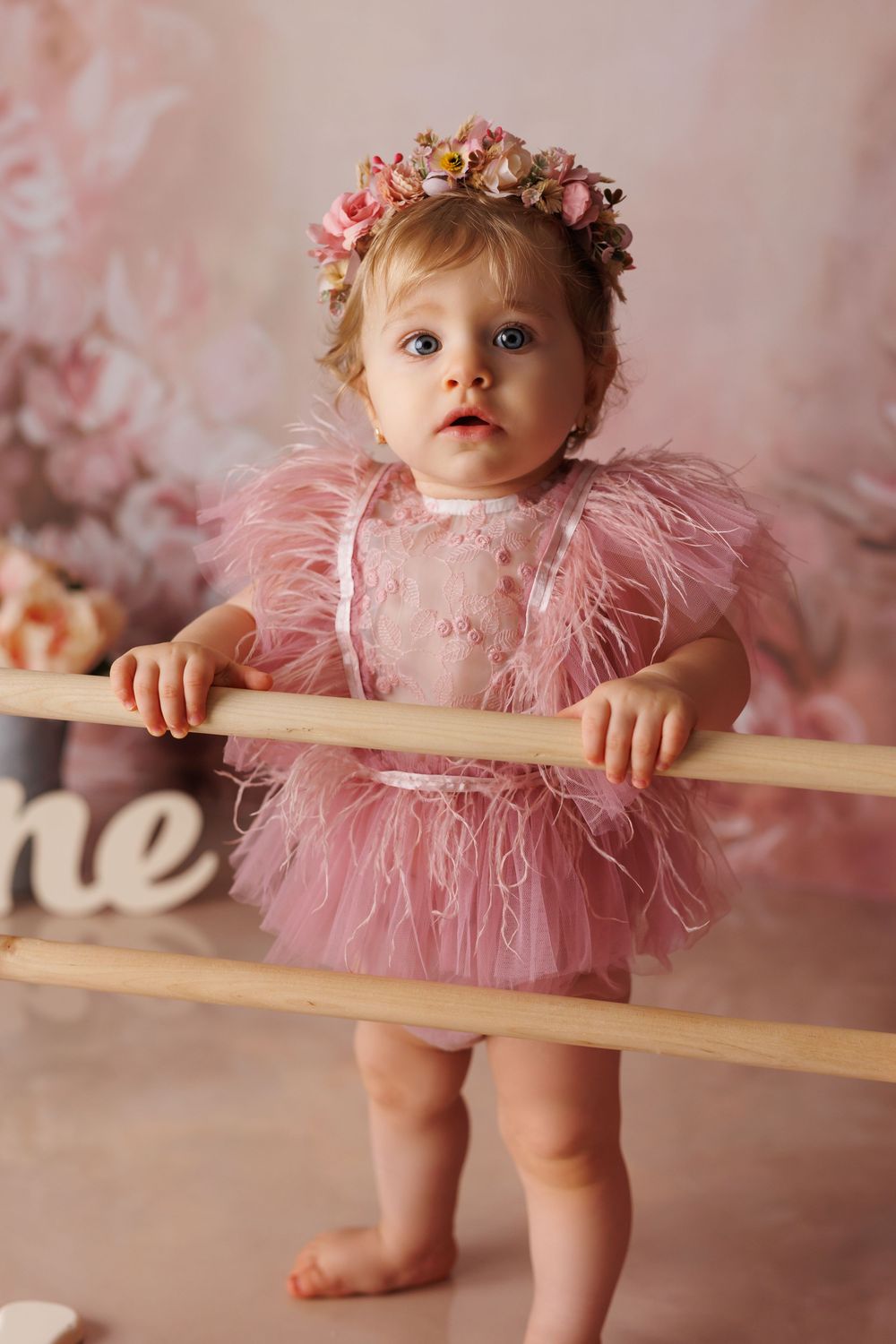 Baby in pink tulle dress and floral crown stands at ballet barre in whimsical photography series.