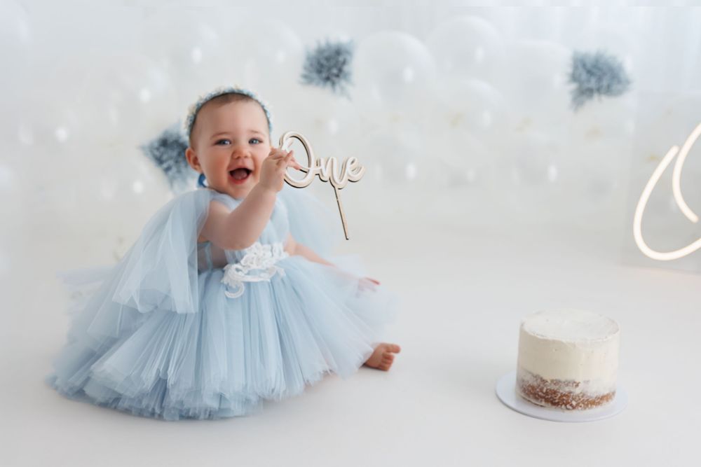 Baby girl in blue tulle dress laughs at first birthday cake smash photo session with balloons.