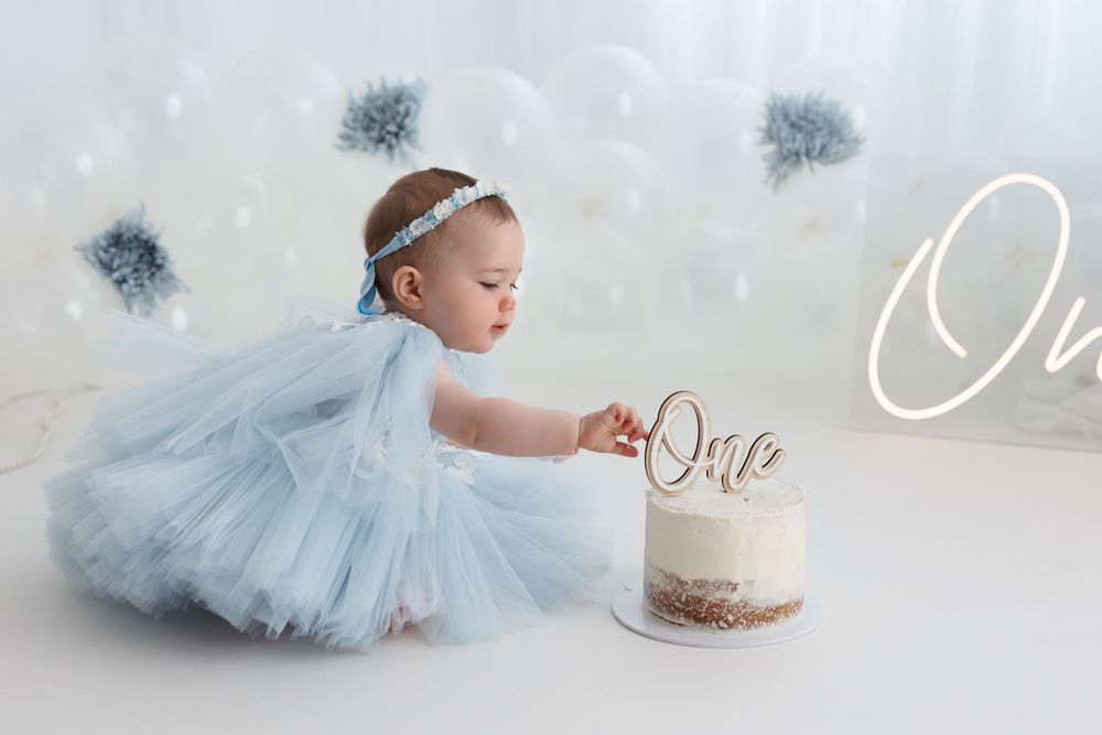 Baby girl in blue tutu dress touches her first birthday cake with a gold 'One' topper at a cake smash session.