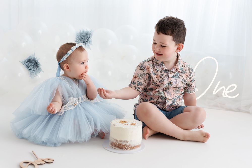 Baby girl in blue tutu dress shares first birthday cake with older brother in a bright studio photo shoot.
