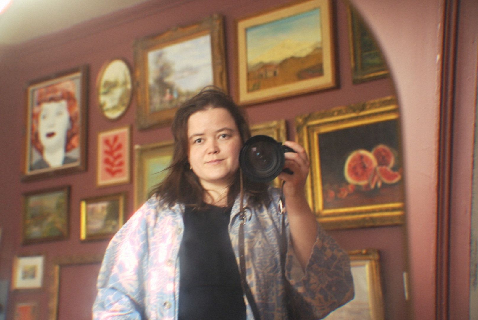 Standing figure wearing floral robe and black shirt in front of wall decorated with colorful framed artwork.