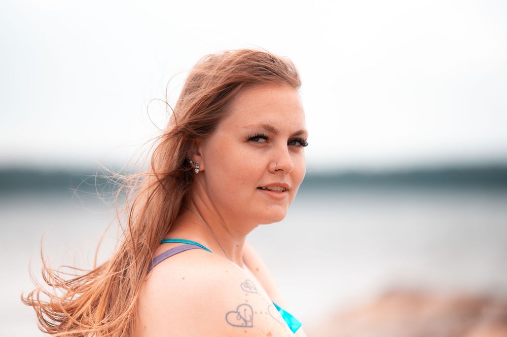 Smiling blonde woman in blue bikini with heart tattoo poses on rocky beach by the sea.