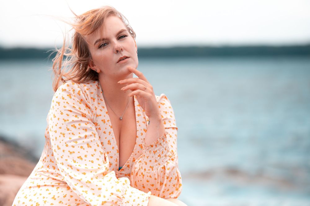 Blonde woman in floral dress sits pensively on rocks by the ocean on overcast day.