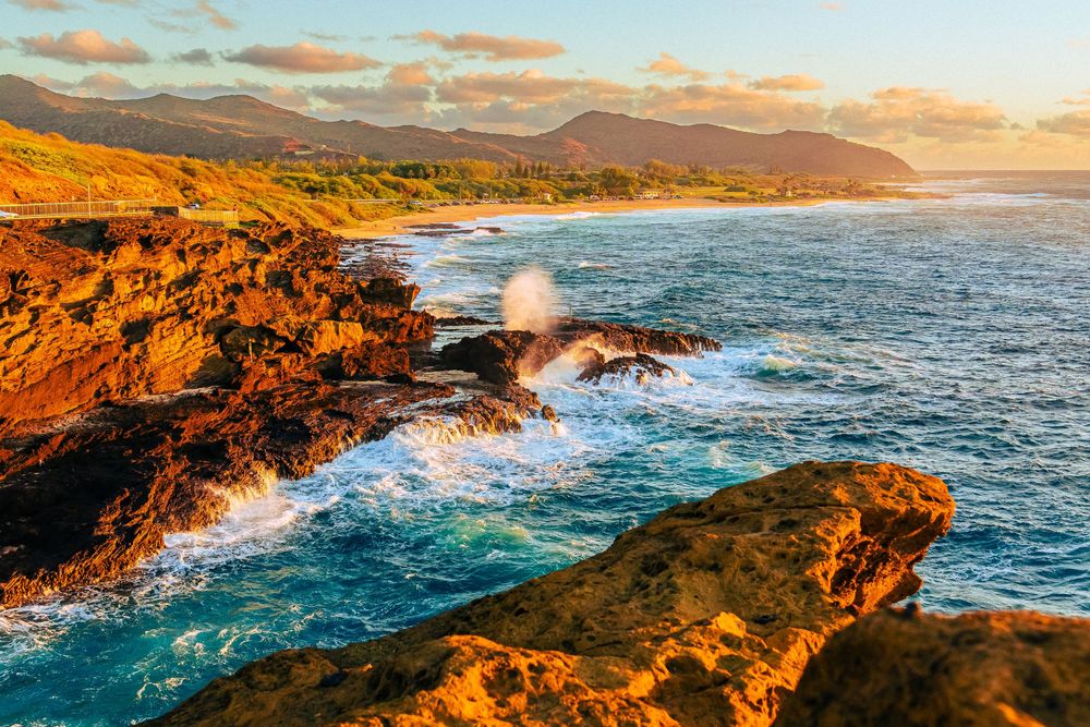 Golden hour light illuminating dramatic rocky coastline with crashing waves and mountain backdrop.