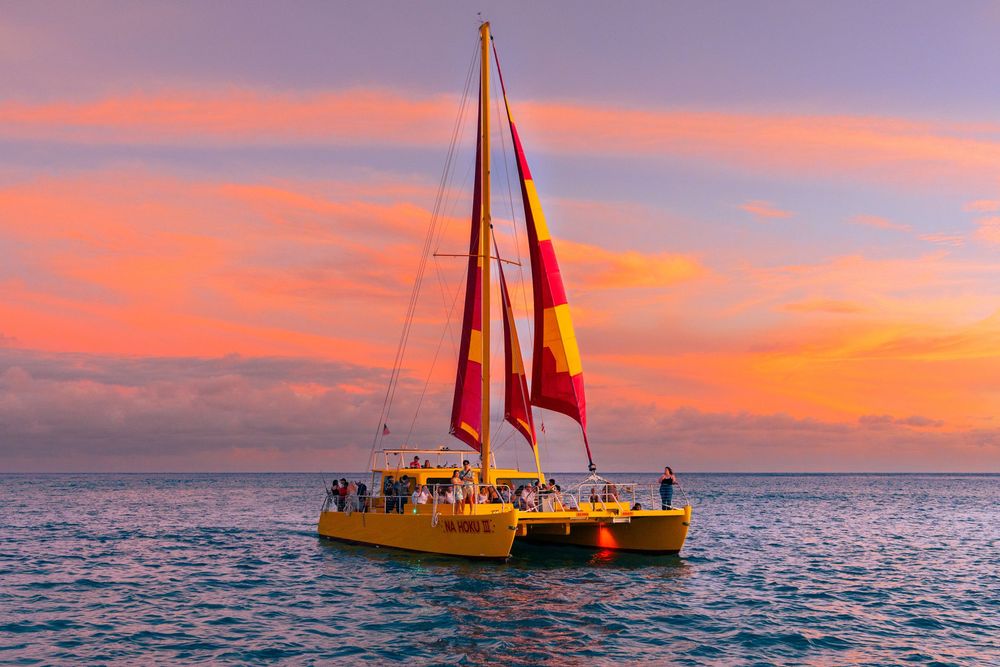 Yellow catamaran with red and yellow sails cruises the ocean at sunset with vibrant pink and orange skies.