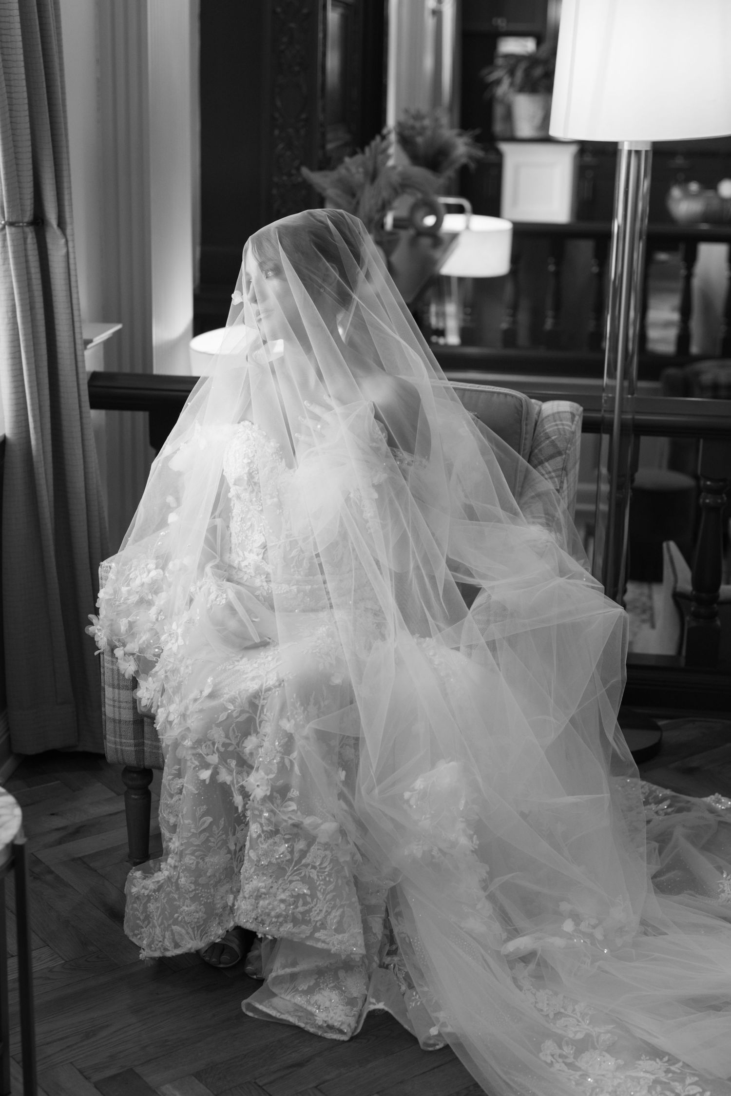 Black and white portrait of bride in lace gown and long veil seated in elegant chair.