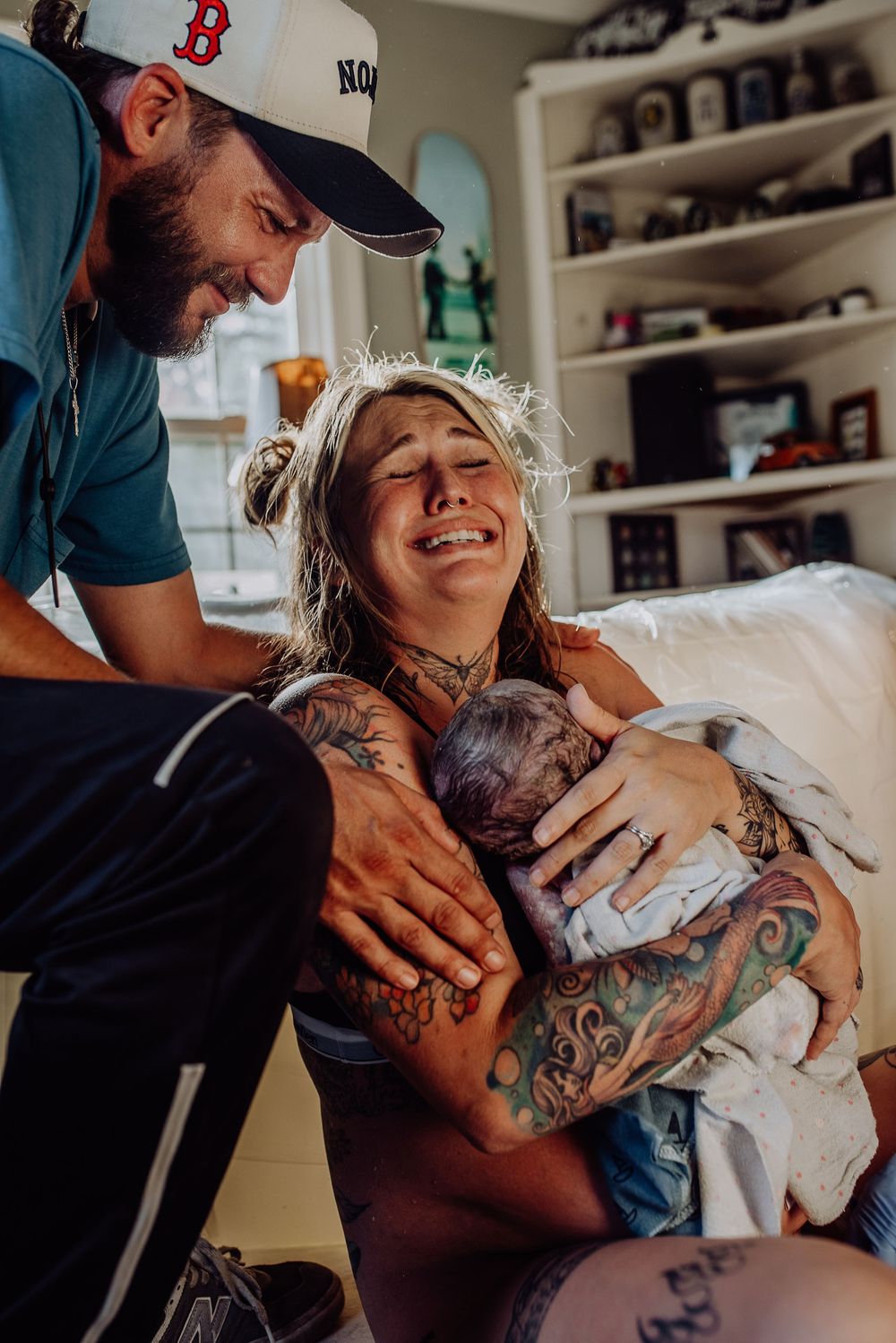 Emotional new mother with tattoos holds newborn baby as tattooed father smiles lovingly beside her in a home setting.