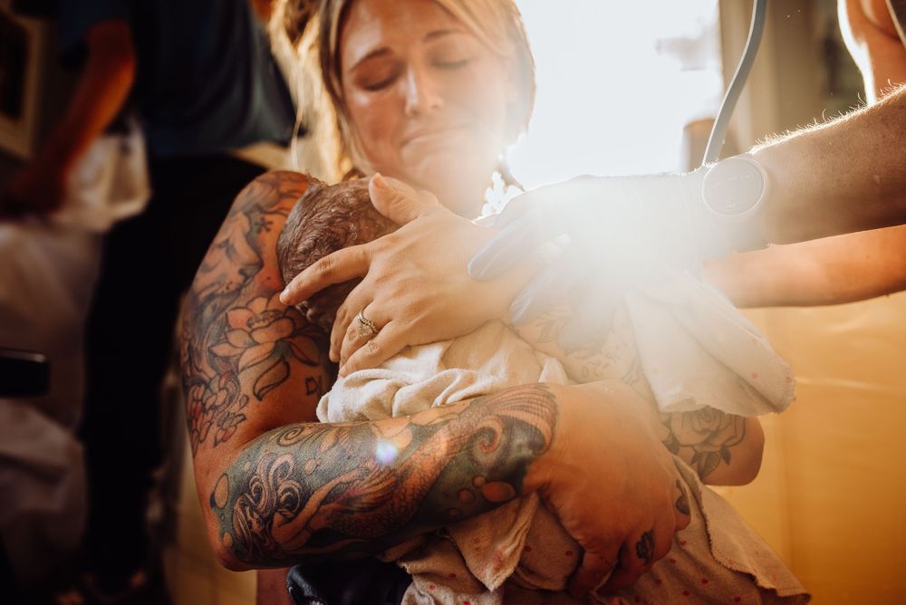Heavily tattooed person cradling a newborn baby wrapped in white cloth in a warmly lit room.