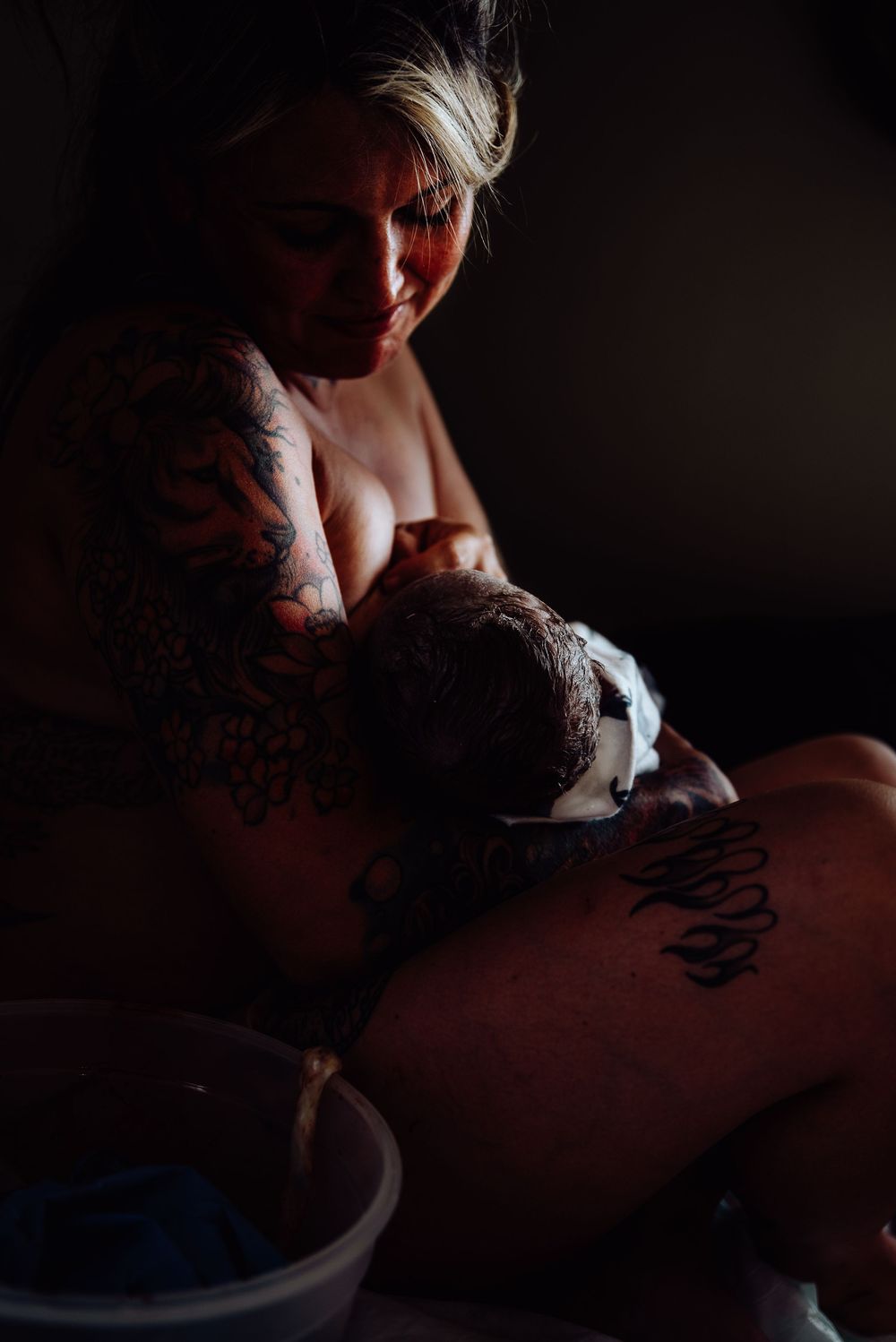 Tattooed mother breastfeeding her newborn baby in a dimly lit room after childbirth.