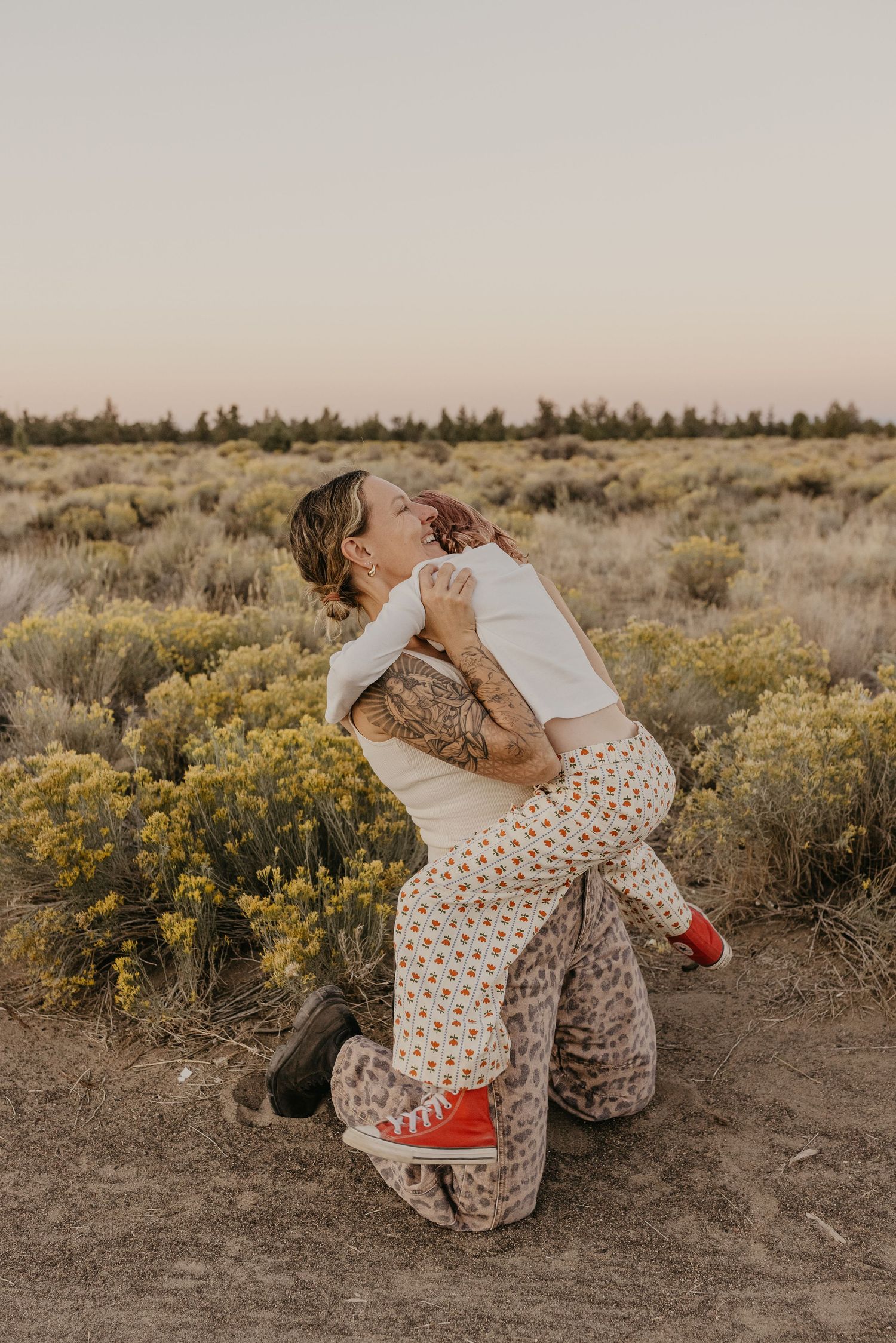 Two women embrace joyfully in a desert sagebrush field at golden hour, one wearing leopard print pants and red sneakers.