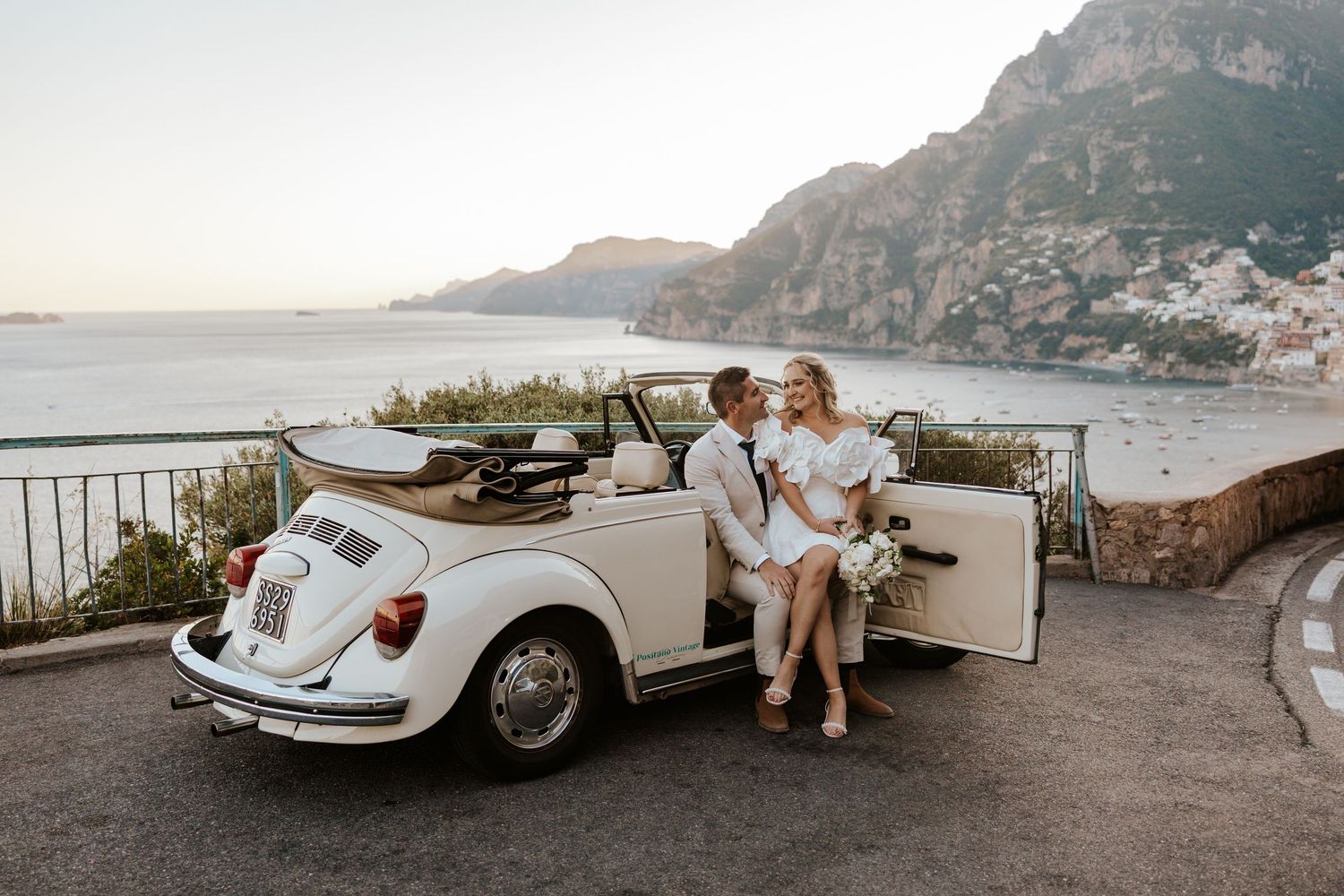 bride and groom leaning on a vintage cream car overlooking Positano in the Amalfi Coast, Italy area