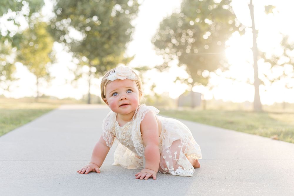 Baby girl crawling along a paved path in a white floral outfit, smiling in warm evening sunlight with trees in the background.