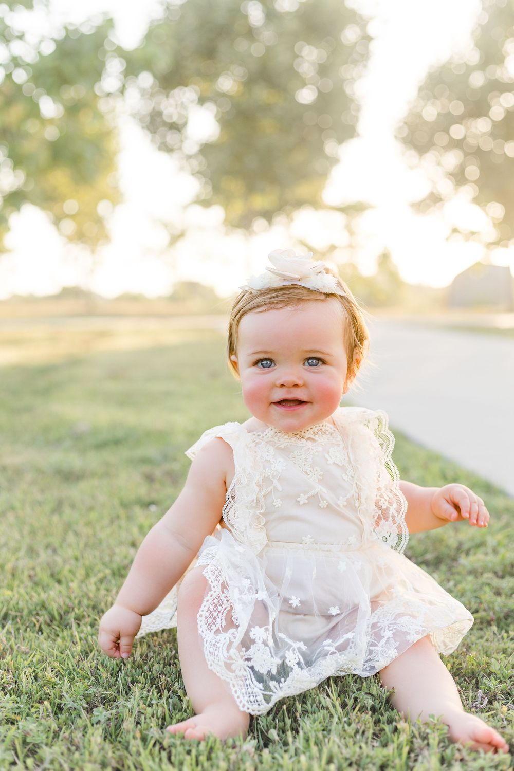 Smiling baby girl sitting in the grass wearing a white floral outfit during a golden-hour outdoor milestone photography session.