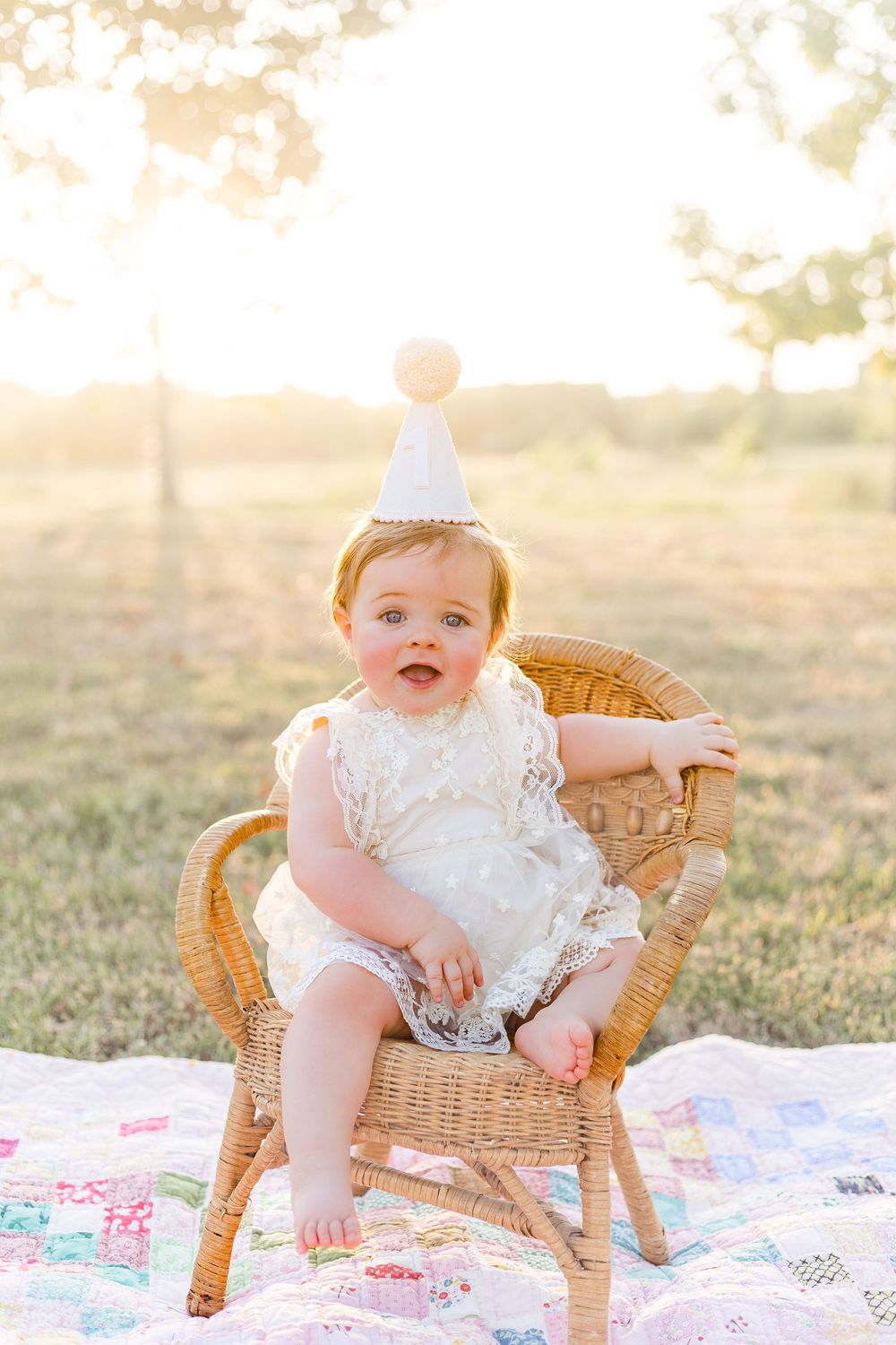 Smiling baby girl wearing a white birthday outfit and party hat while sitting on a blanket outdoors in warm evening sunlight.