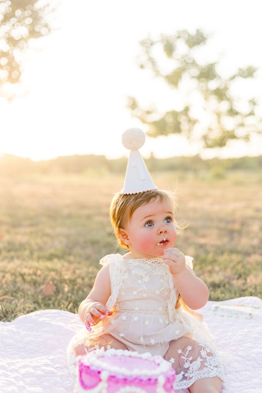 Baby girl wearing a white birthday outfit and party hat sitting beside a pink cake during an outdoor first birthday photo session.