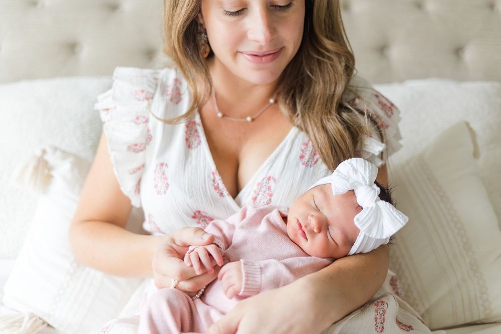 Mother smiling down at her newborn baby dressed in pink while cuddling together on a soft bed during a lifestyle newborn session.