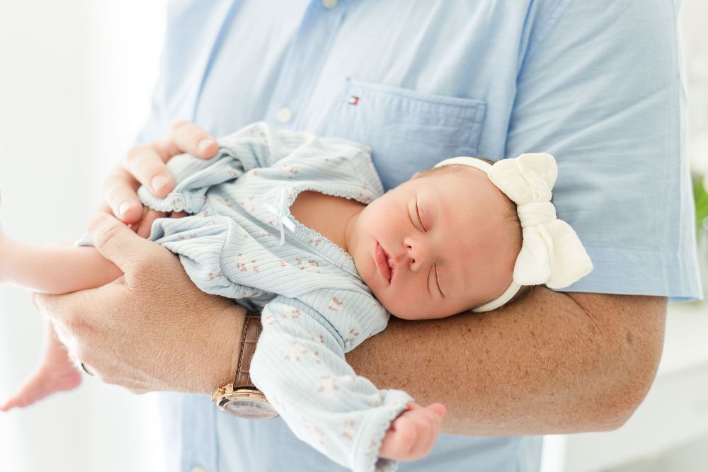 Sleeping newborn baby wearing a pale blue outfit and white bow headband, gently cradled in a parent's arms.