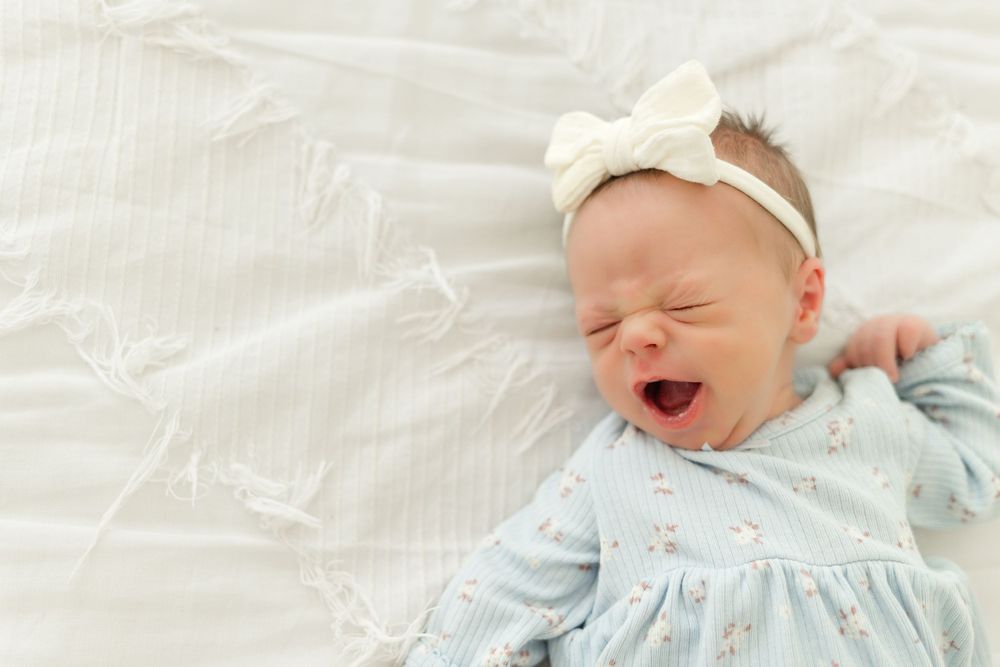 Baby girl wearing a pale blue outfit and white bow headband, yawning while lying on soft white bedding.