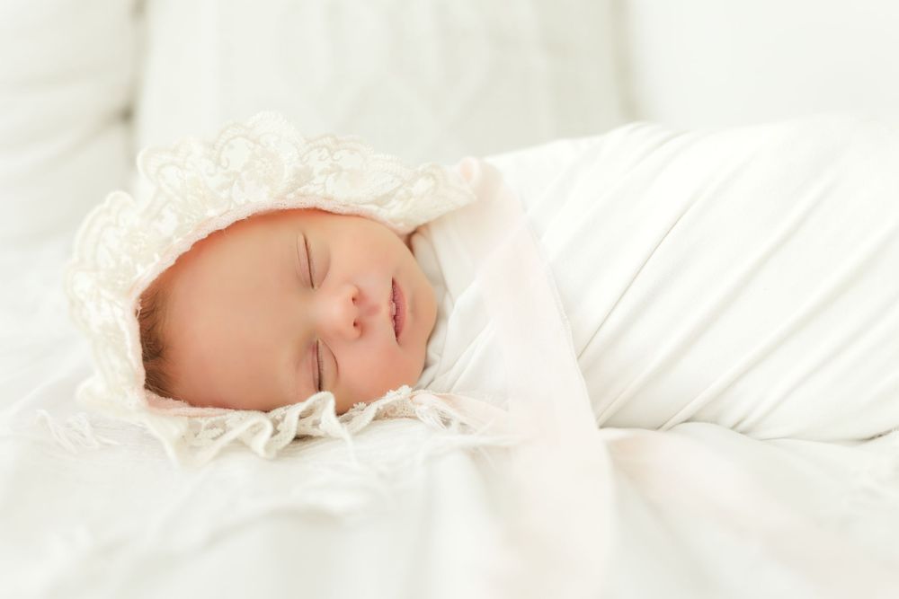 Close-up portrait of a peacefully sleeping newborn baby wrapped in white fabric and wearing a delicate lace bonnet.