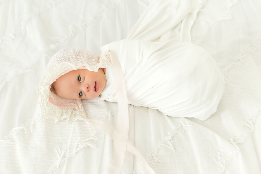 Newborn baby lying on soft white bedding, wrapped in a white blanket and wearing a delicate bonnet during a studio portrait session.