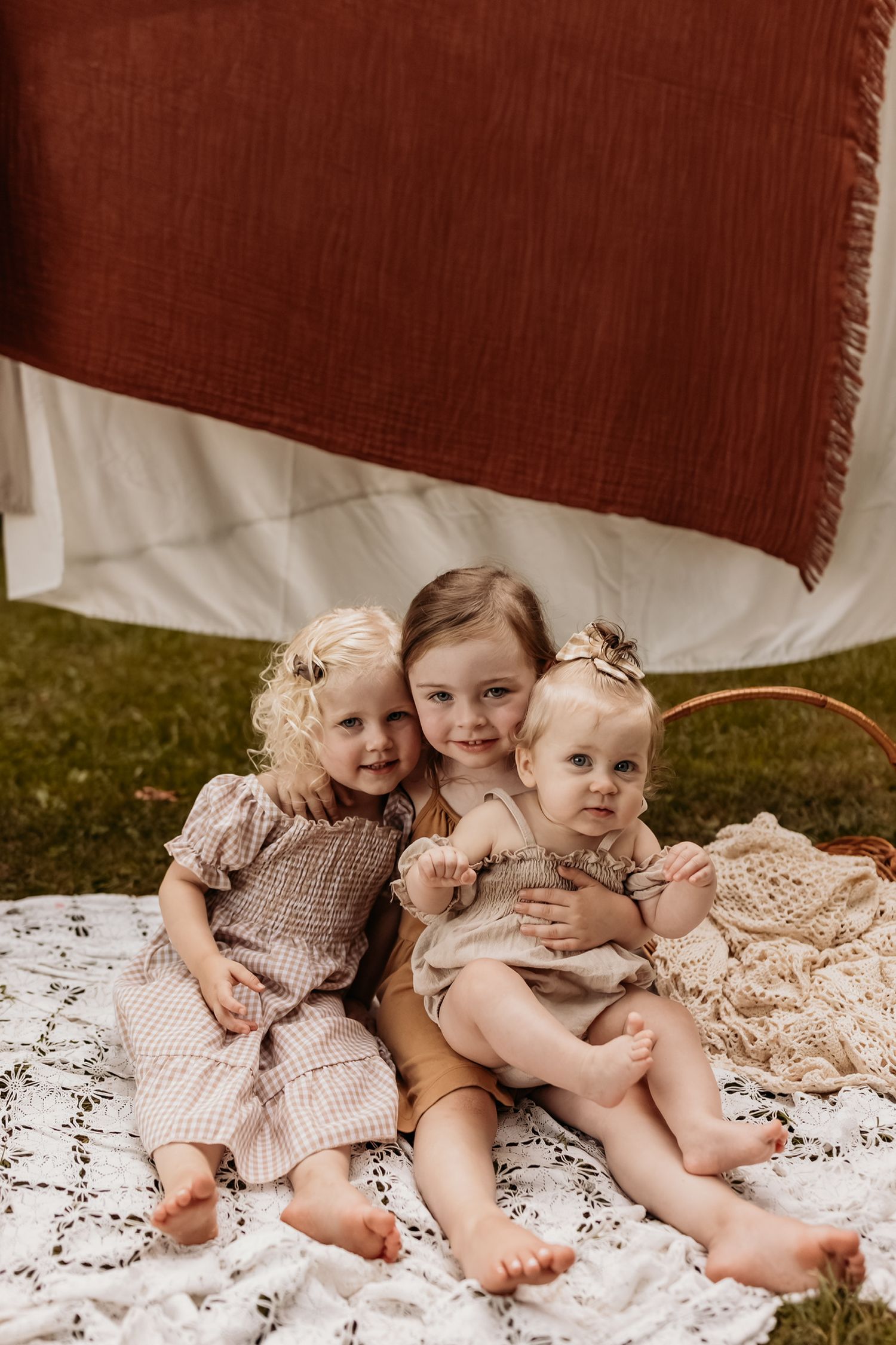 Three children sitting together on white lace blanket under rustic hanging textile.